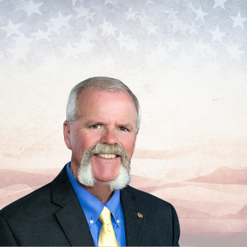 Man in a suit, blue shirt, and yellow tie with a large mustache against a backdrop of American flags.