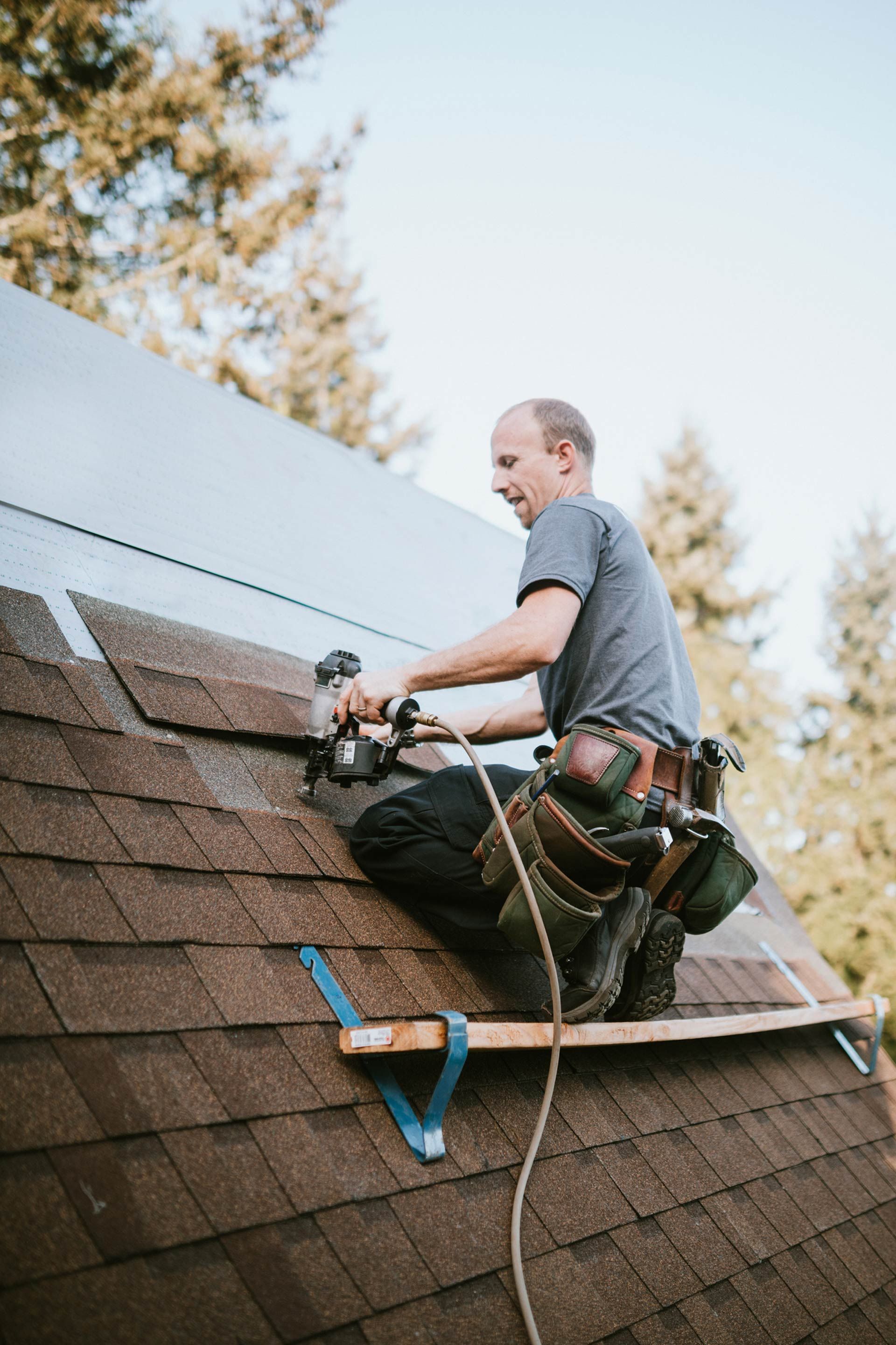 Roofer in gray shirt, securing shingles with a nail gun, working on a brown shingled roof outdoors.