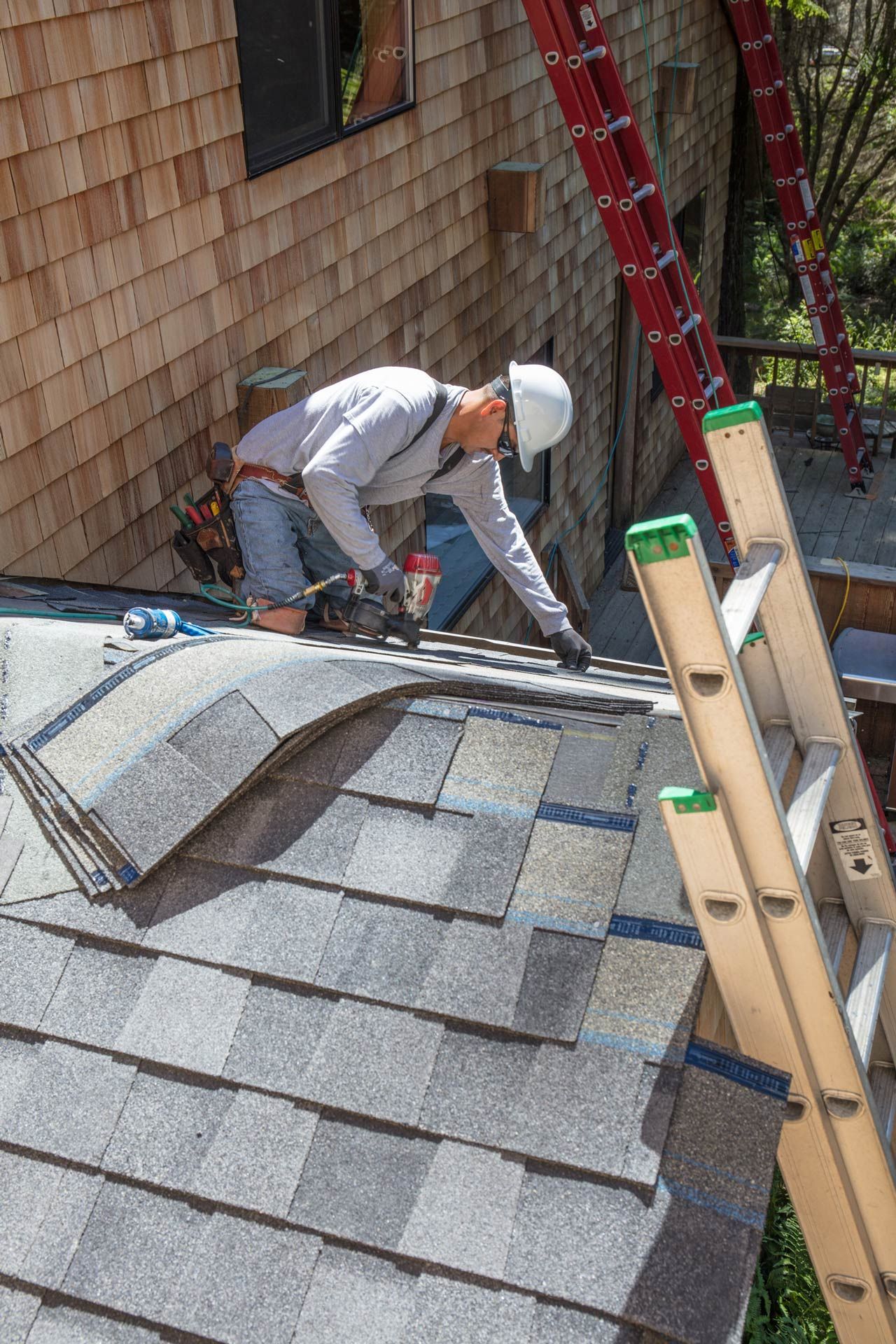 A construction worker, wearing a hard hat, works on a shingle roof. Ladders and a partially shingled wall are visible.