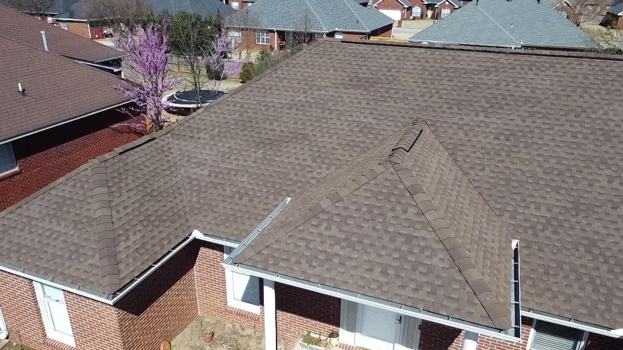Brown shingled roof on a brick house, viewed from above on a sunny day. Other houses and trees are in the background.