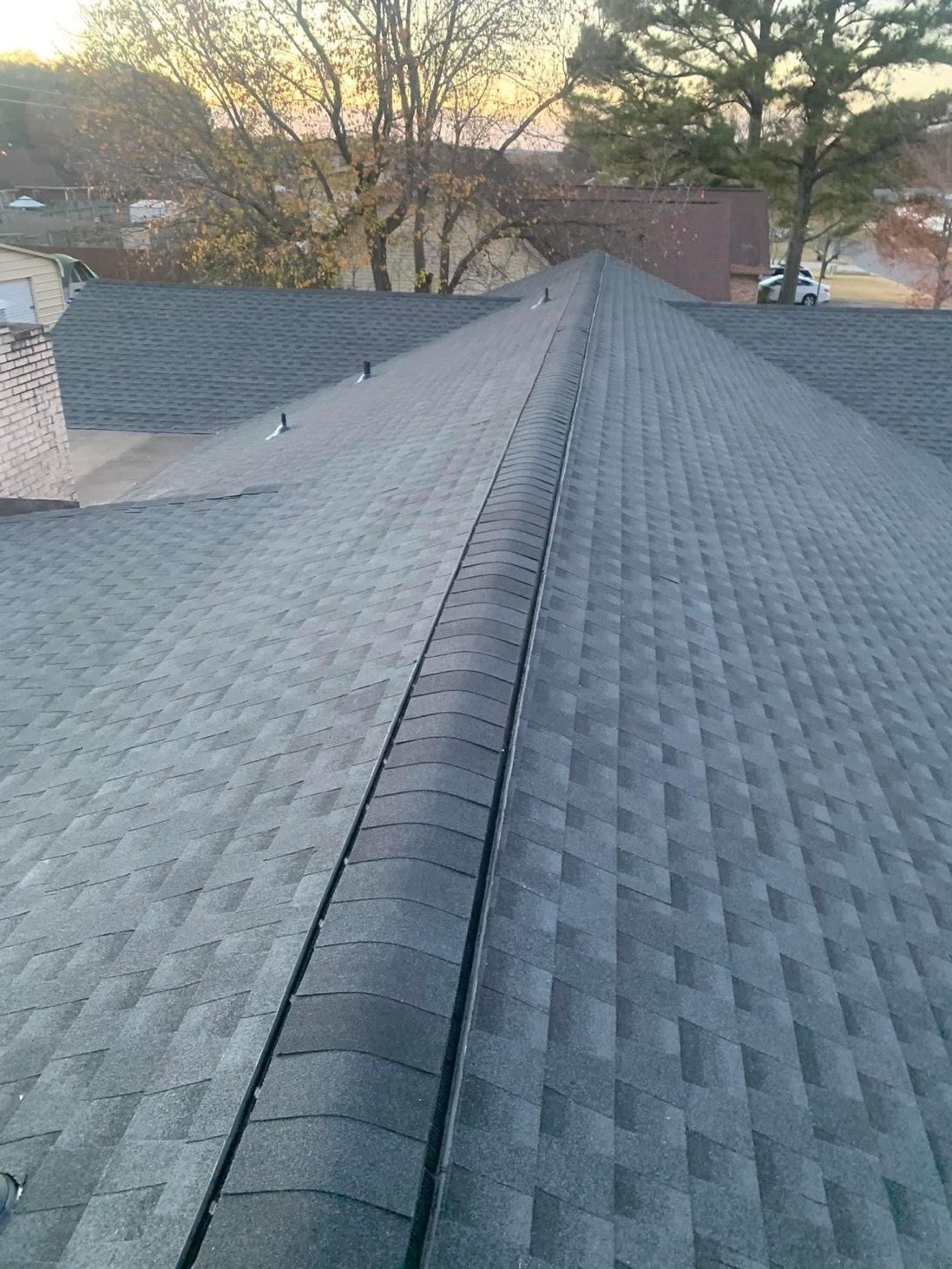 A rooftop covered in gray asphalt shingles, with a central ridge and buildings and trees in the background.