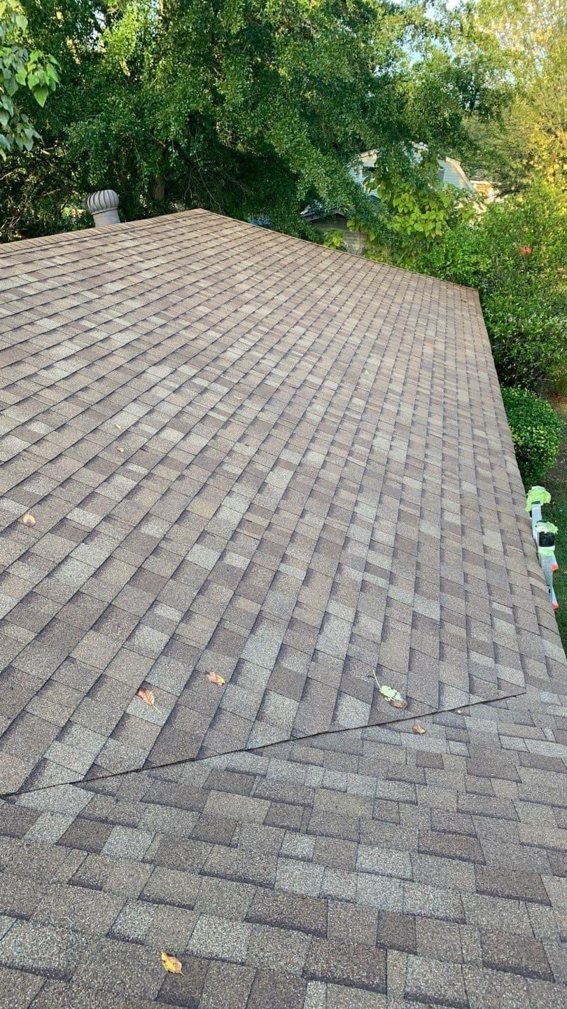 A rooftop covered in brown asphalt shingles, viewed from above. Green trees are visible in the background.
