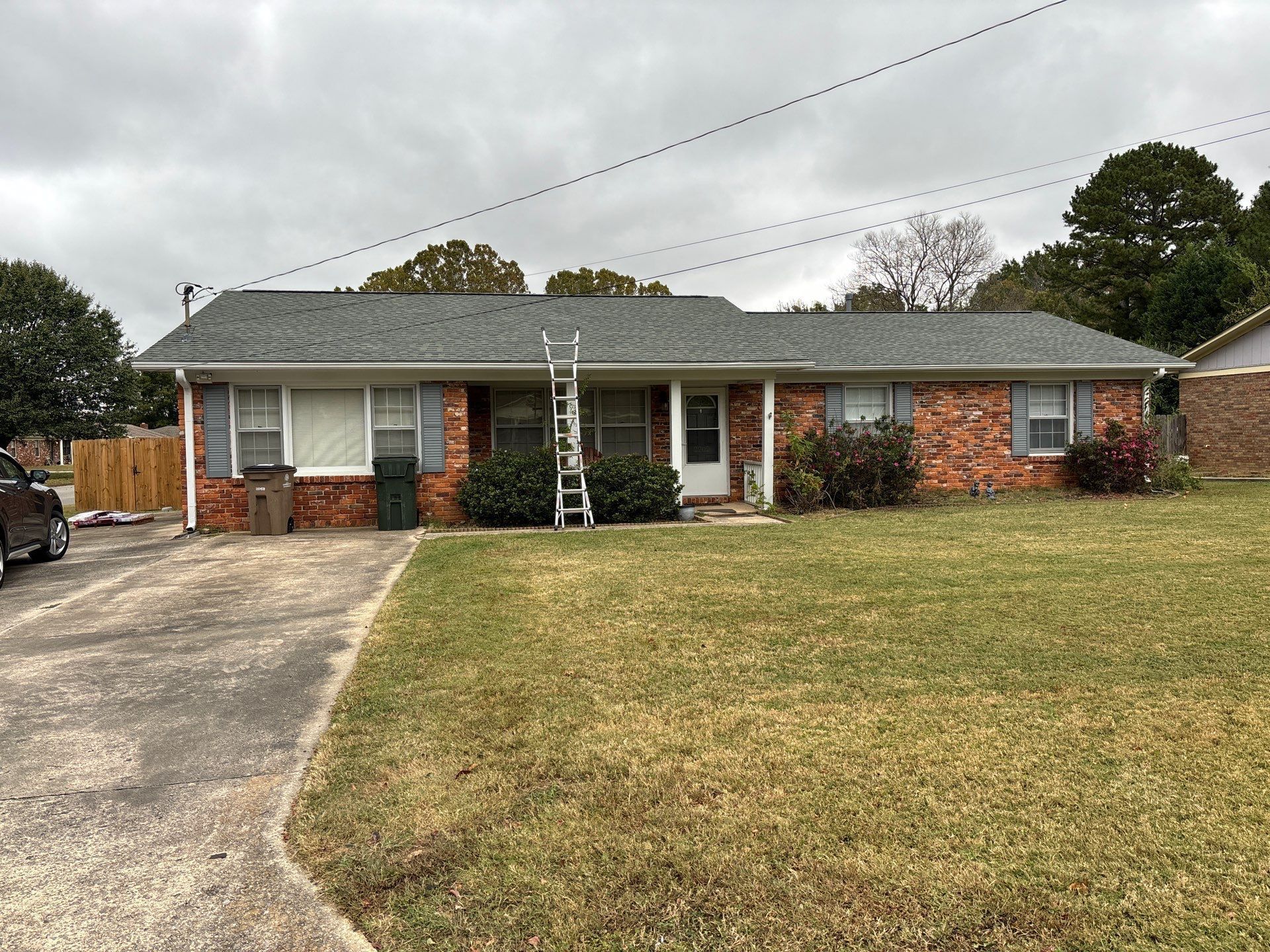 Brick ranch house with a gray roof and a ladder leaning against it on a cloudy day. A lawn is in the foreground with a driveway to the left.