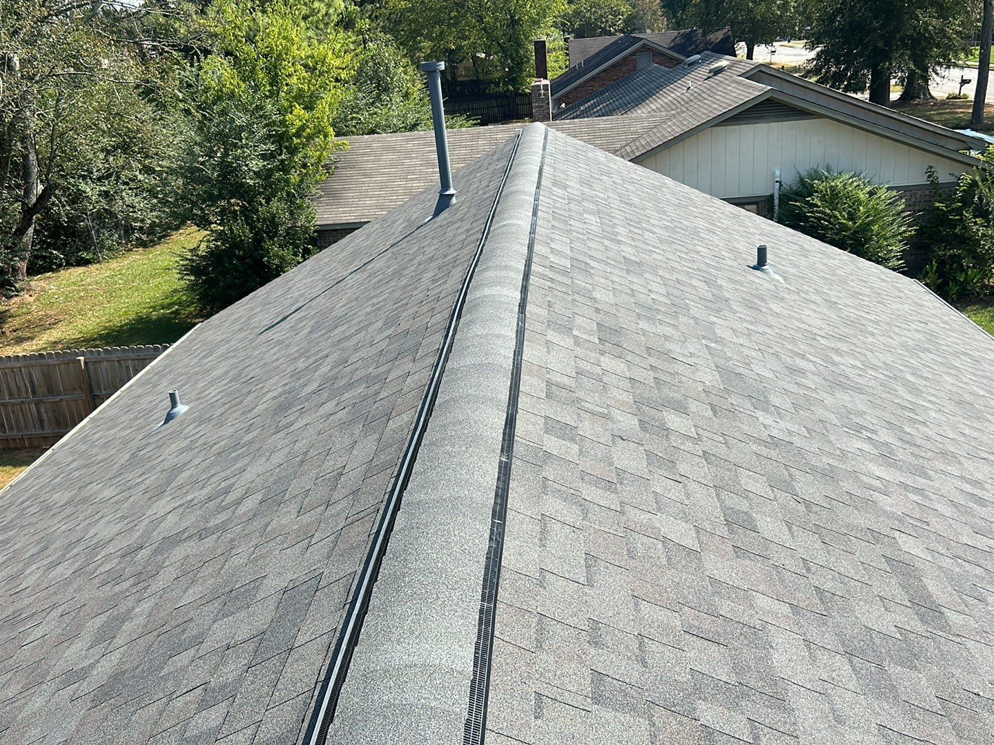 View of an asphalt shingle roof, likely on a house, with a central ridge and vents. Green trees and other houses are in the background.
