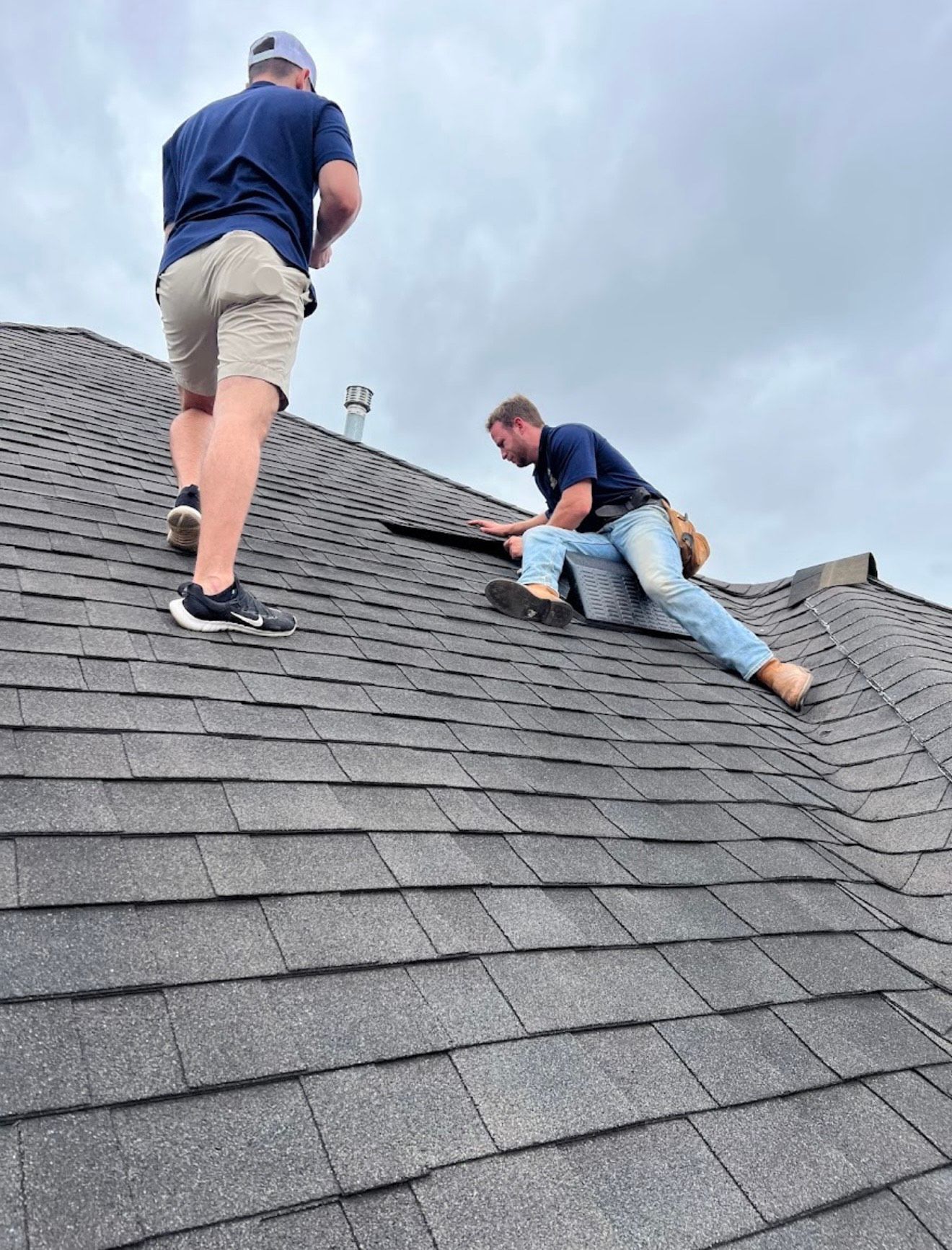 A roofer kneels on a shingled roof, using a nail gun. Another person's legs are visible to the left.
