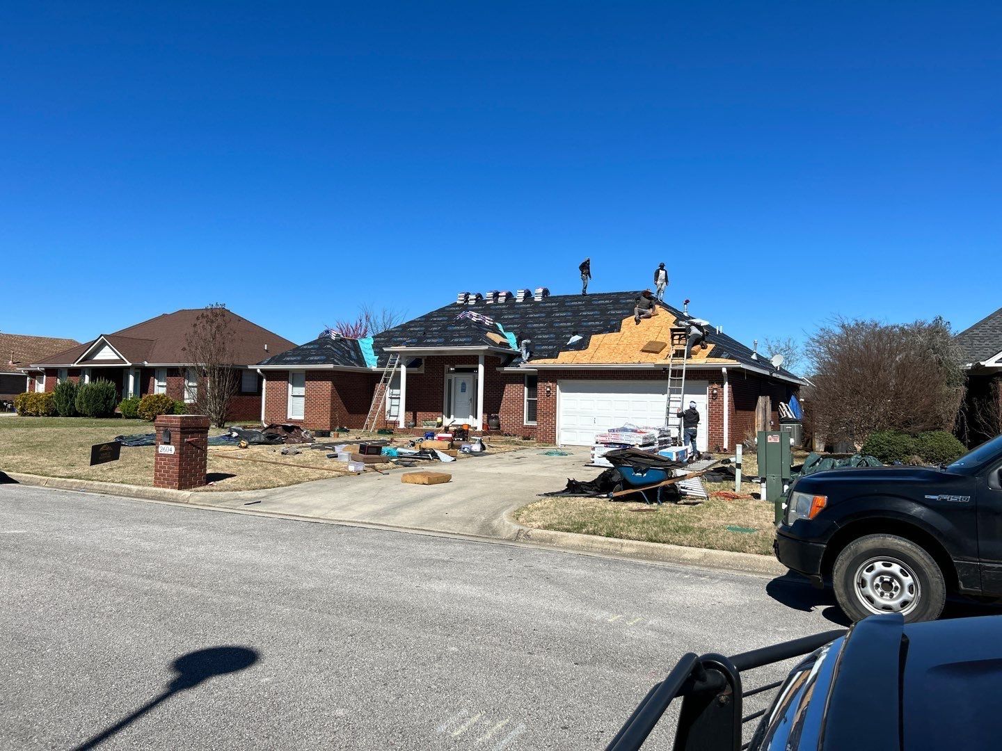 Two roofers installing shingles on a house roof; a chimney stands on the roof, and trees are visible in the background.