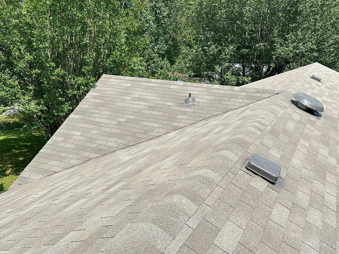 An overhead view of a roof with shingles, a vent, and trees in the background.