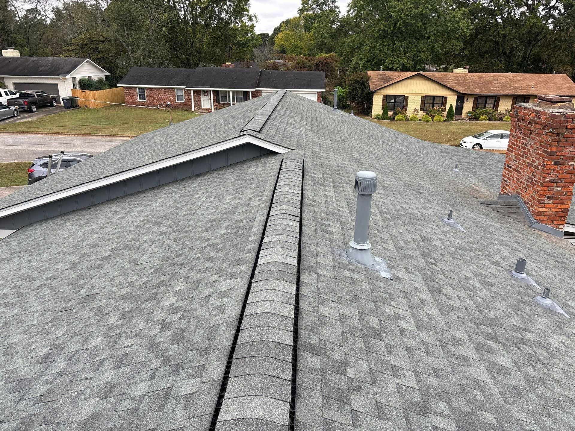 View of a residential roof with gray shingles, a central ridge, and a chimney in a suburban setting.