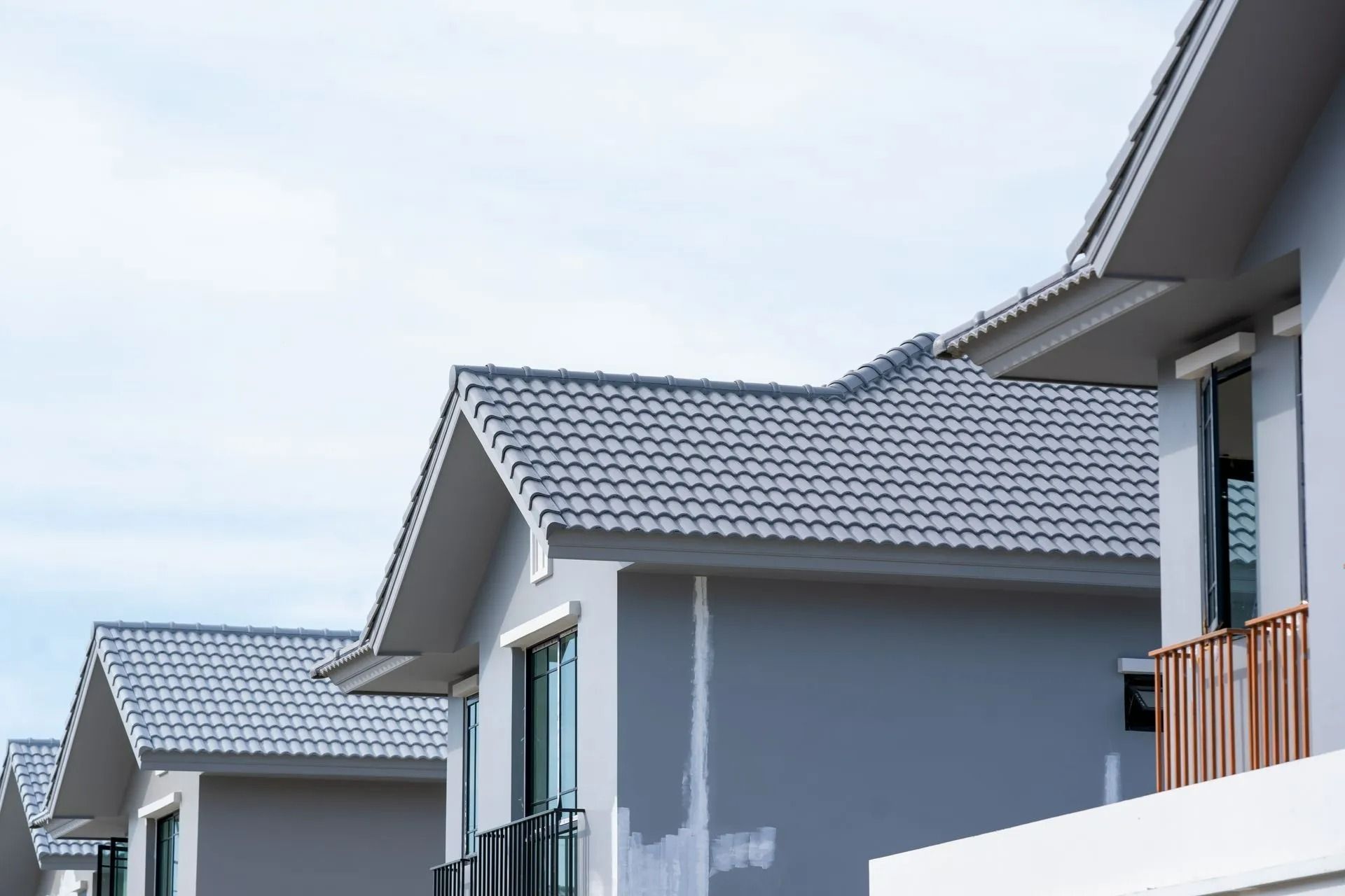 Row of gray houses with matching tiled roofs against a light blue sky. Balconies visible on some units.