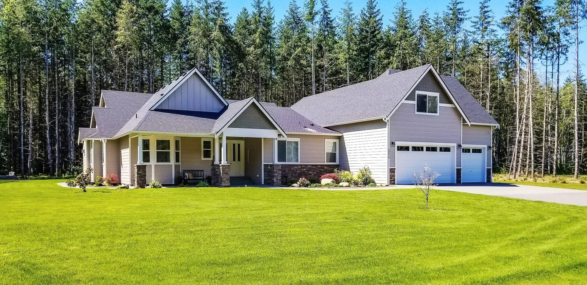 A large house with a light-colored exterior, surrounded by green grass and a backdrop of tall trees.