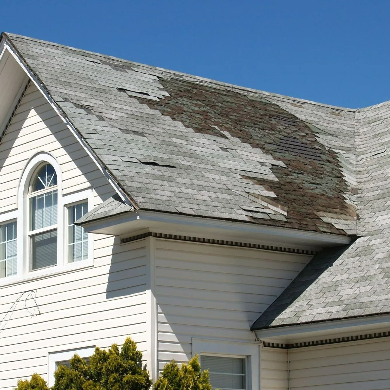 A house with significant roof damage; torn and missing shingles are visible. White siding, arched window, and blue sky are also seen.