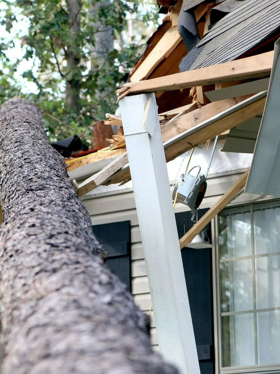 A large tree has fallen, crashing into the roof and porch of a house, causing significant damage.