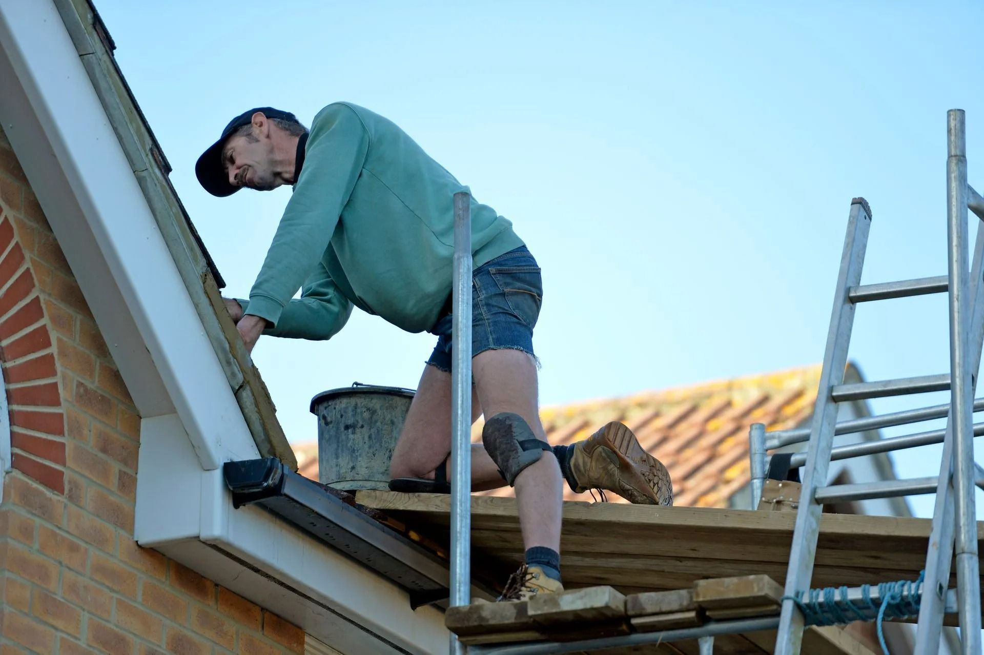 Man working on a roof, kneeling on a scaffold and installing trim. Wearing green shirt, shorts, and a cap; brick building, clear sky.