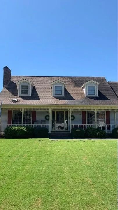 A two-story house with a large green lawn under a clear blue sky. The house has a porch, red shutters, and three dormer windows.