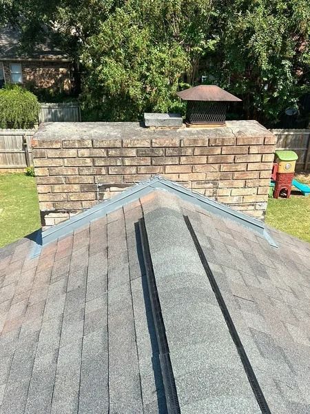 Brick chimney on a shingled roof, viewed from above. The chimney has a metal cap; surrounding is a green yard and trees.