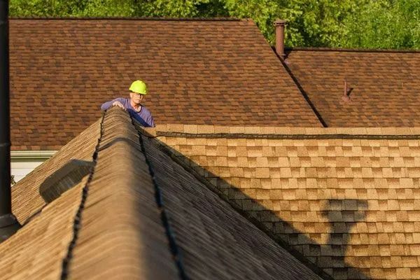 Roofer in a yellow hard hat working on a brown shingled roof on a sunny day.