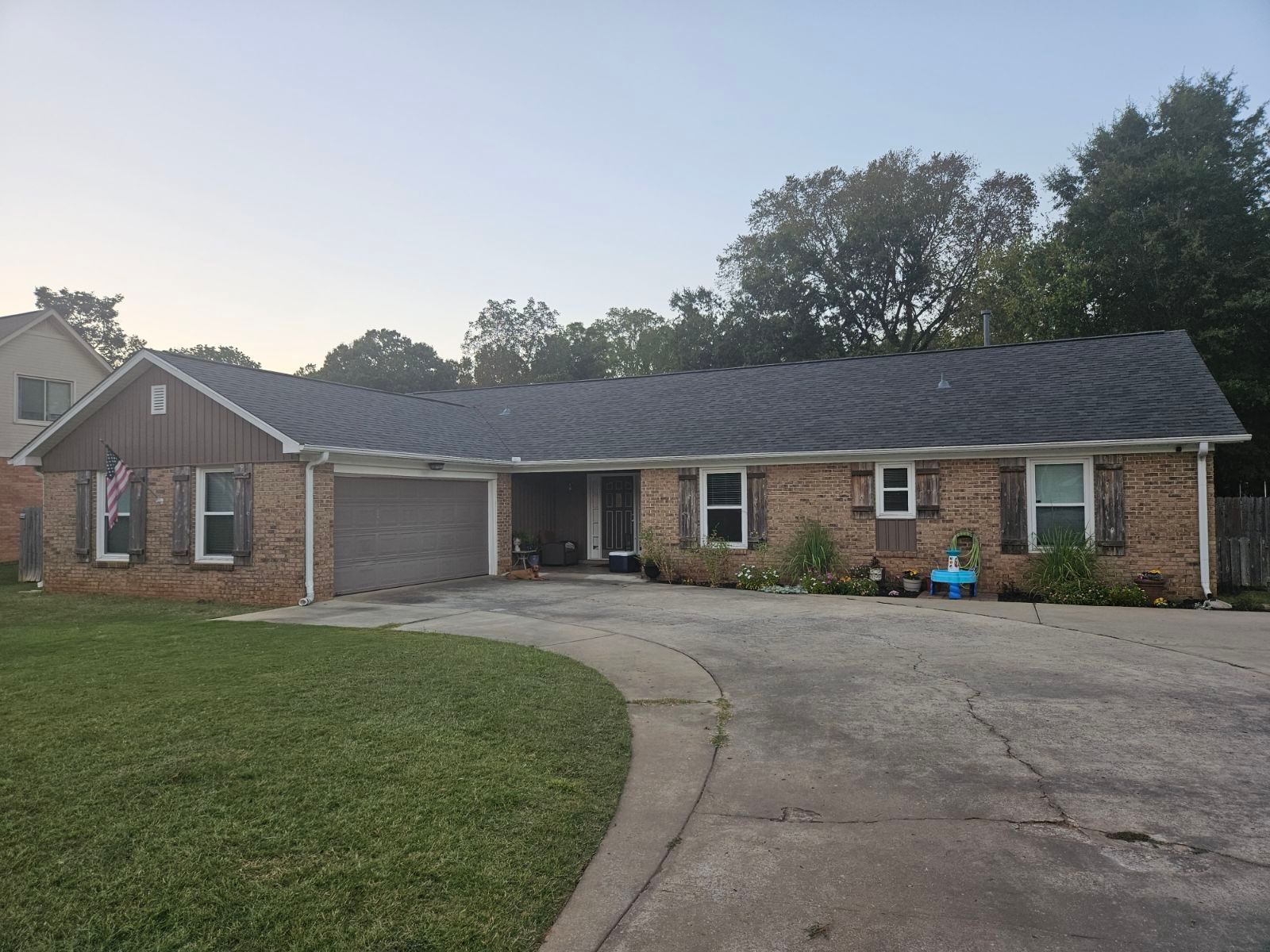 Brick ranch-style house with a driveway, green lawn, and trees. The house has a gray garage door and shuttered windows.