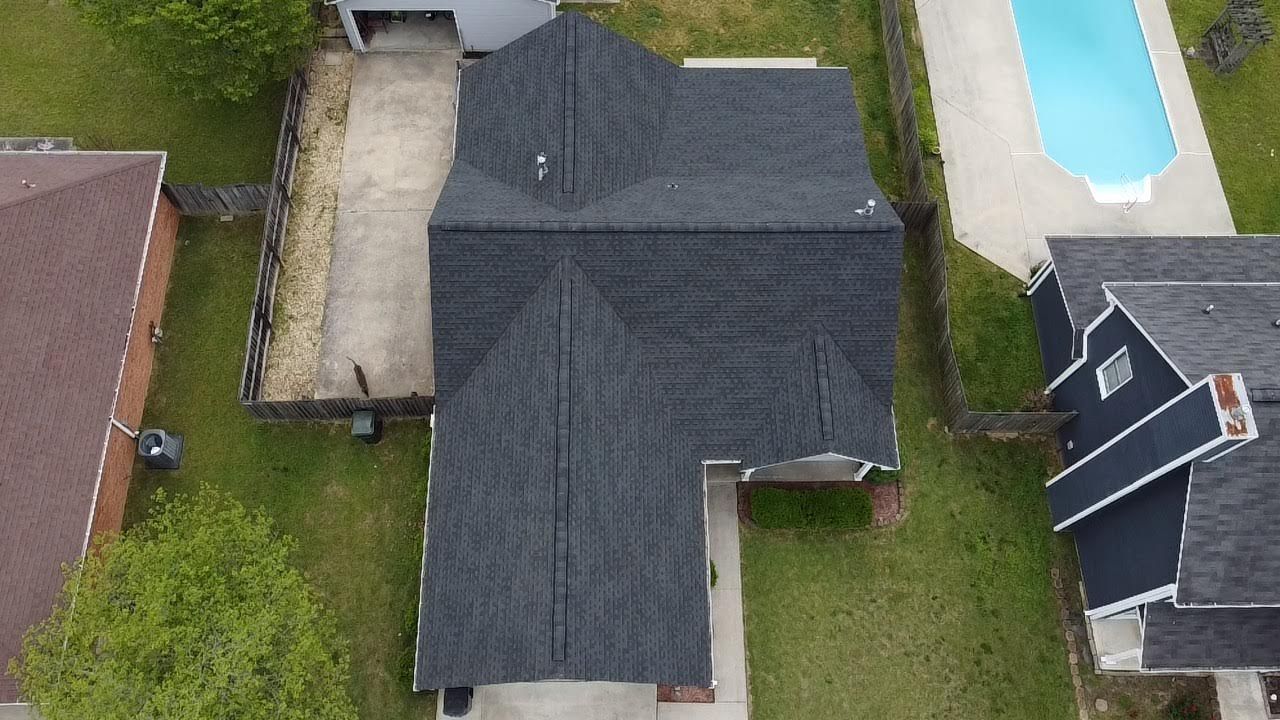 Overhead view of a house with a dark gray roof, a driveway, and a swimming pool. Green grass surrounds the house.