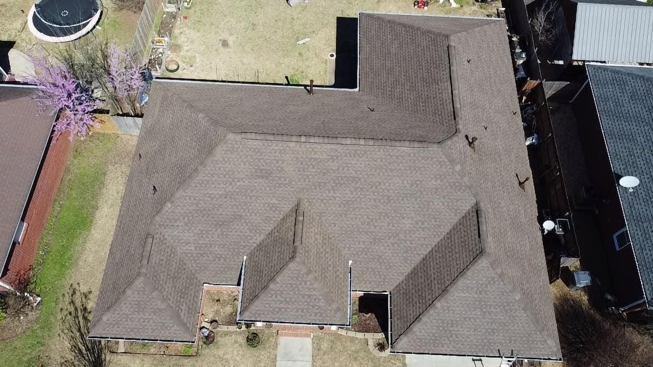 Brown shingled roof of a house, seen from above, with a circular feature and another house to the side.