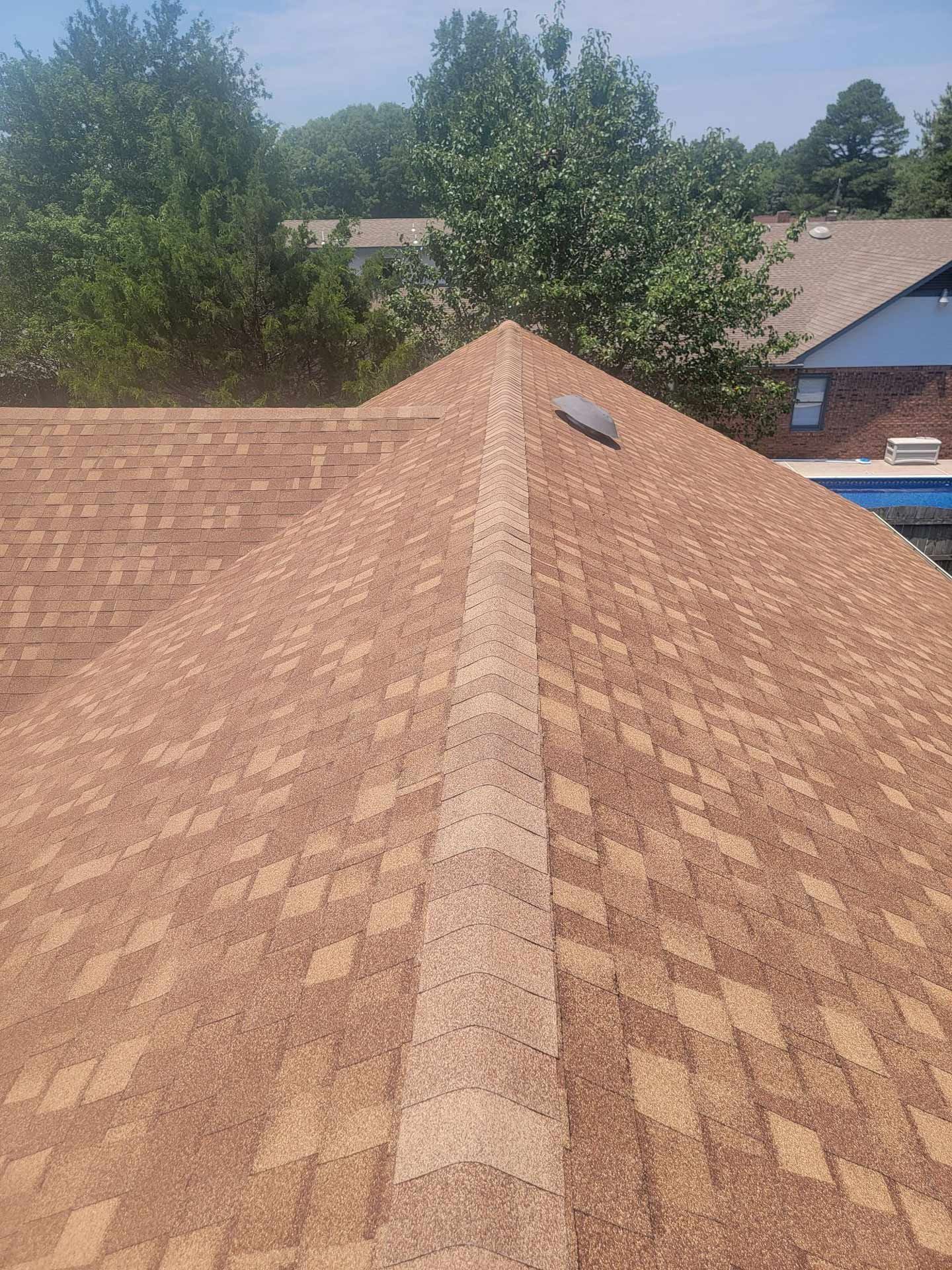 Brown shingle roof with a light-colored ridge, seen from above. Trees and part of a building with a pool are visible.