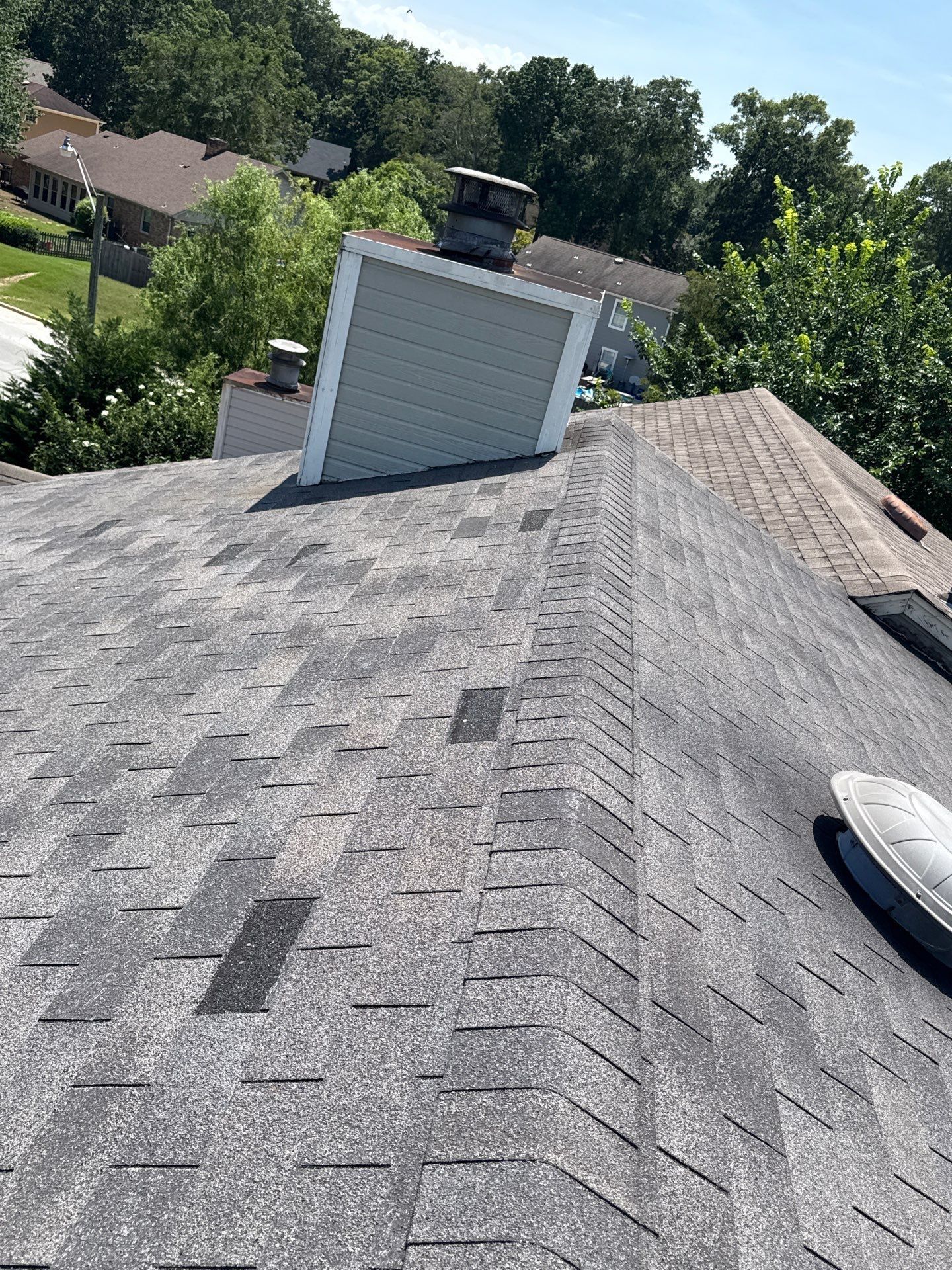 Man in hard hat on a ladder inspecting the roof and window trim of a tan house. He is holding a device and looking upward.