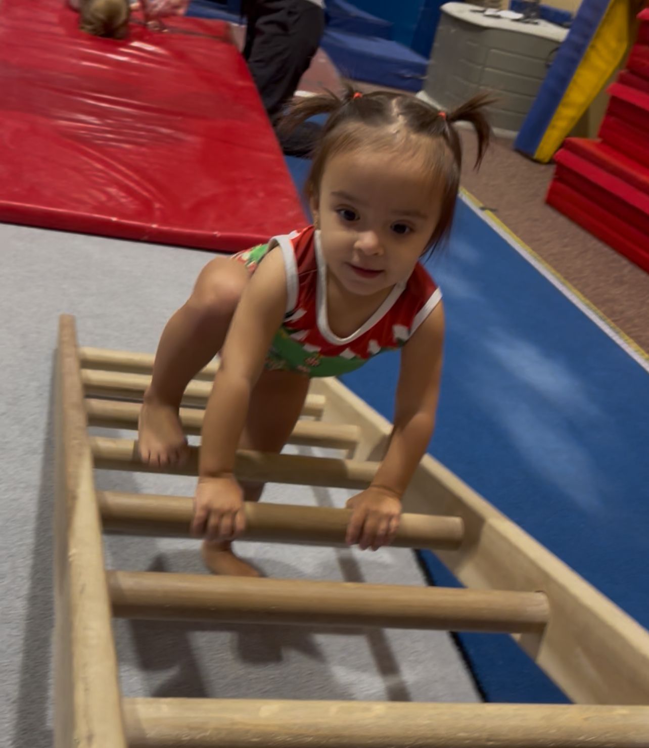 Toddler in a gymnastics leotard climbing a wooden ladder in a gym.