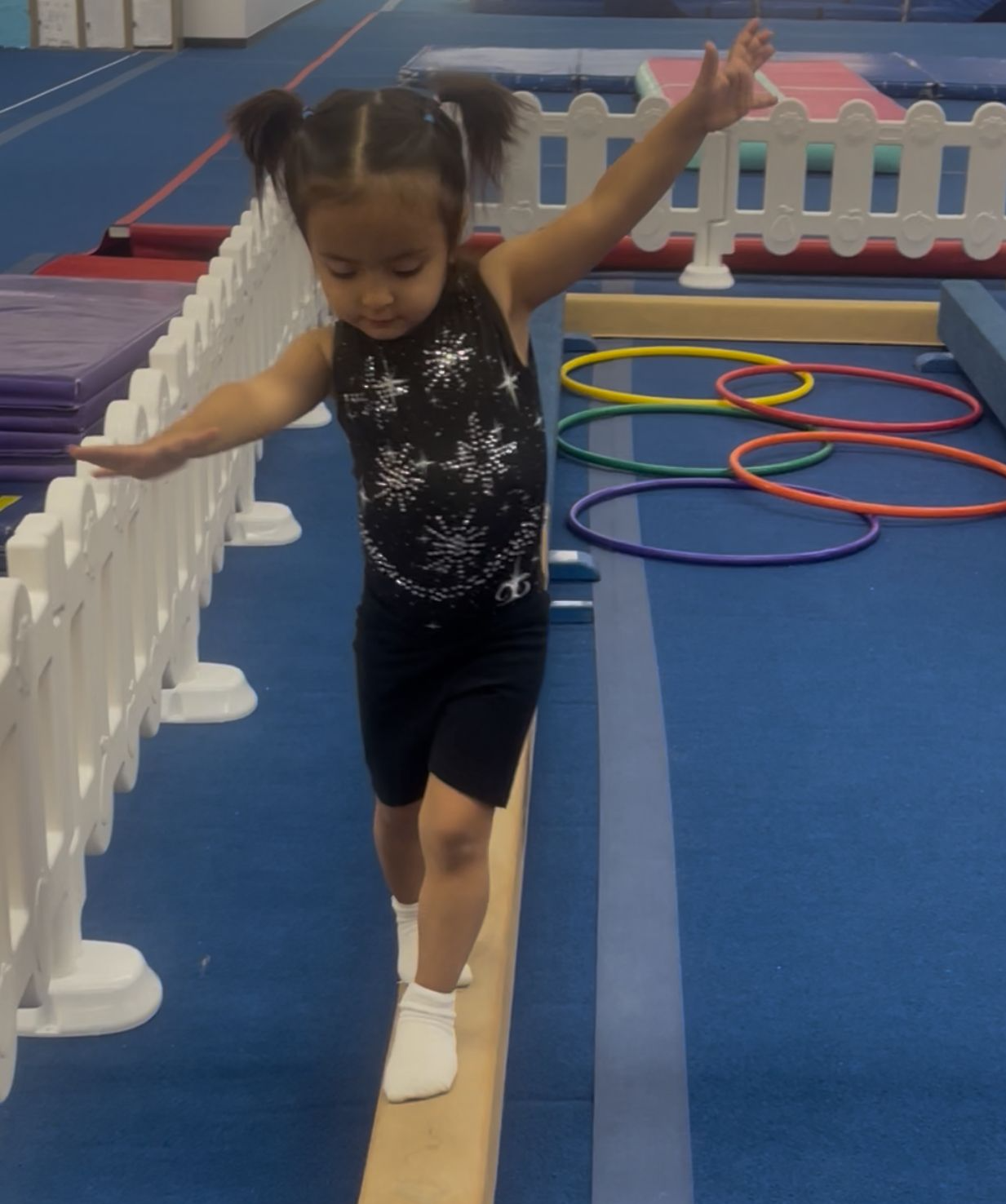 Young child balances on a beam, arms out, in a gym.