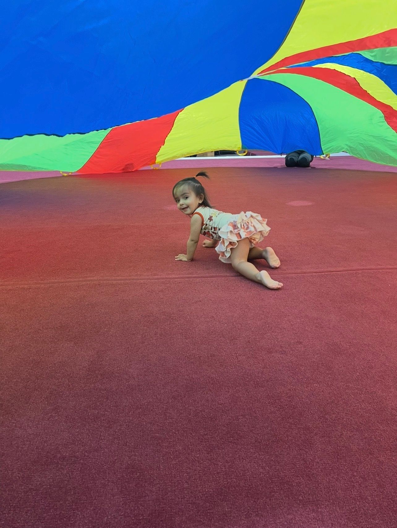 Young child crawling on red surface under a colorful parachute, smiling.