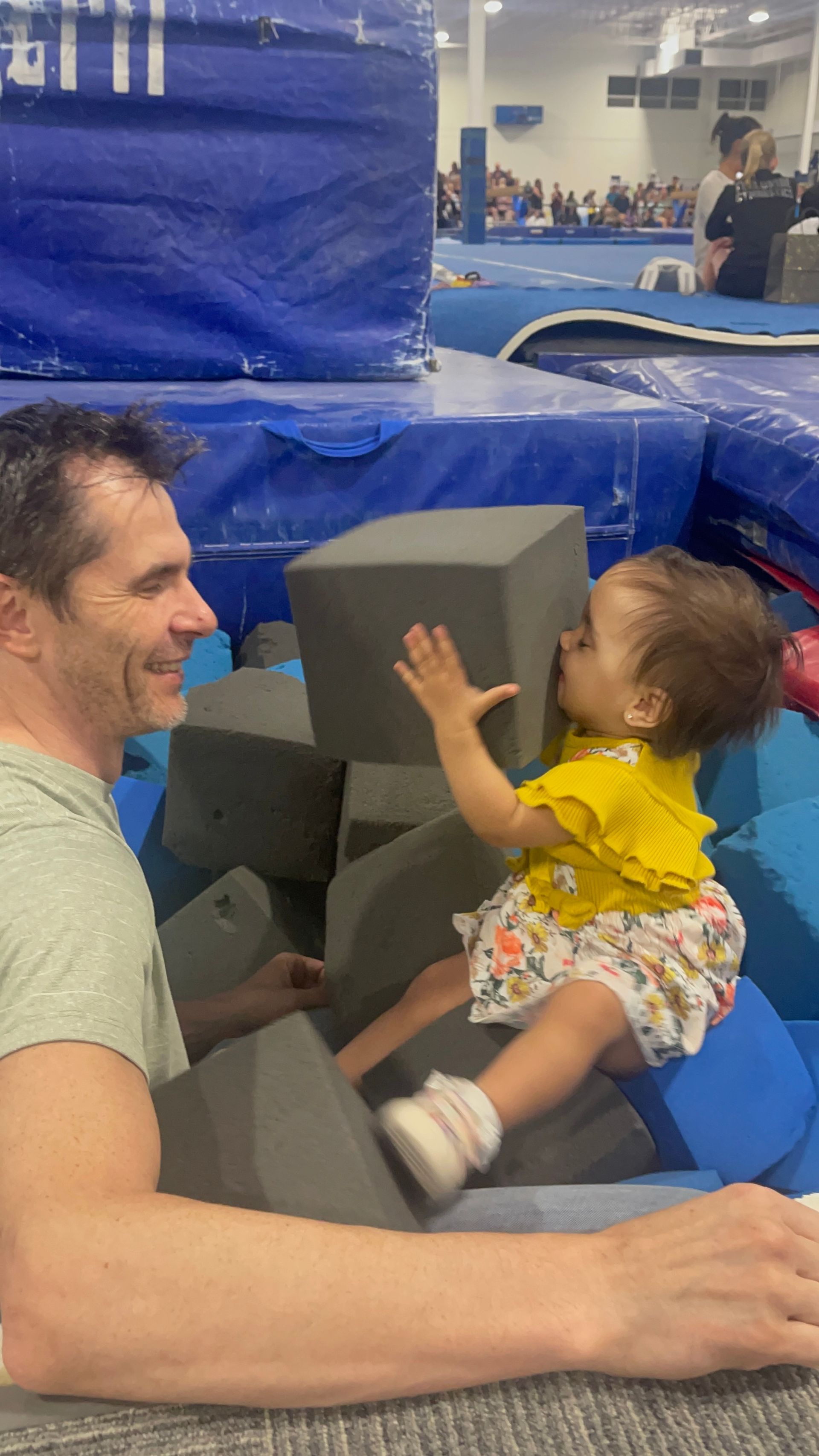Man smiles at a toddler in a foam pit, who reaches for a block. The setting appears to be a gym.