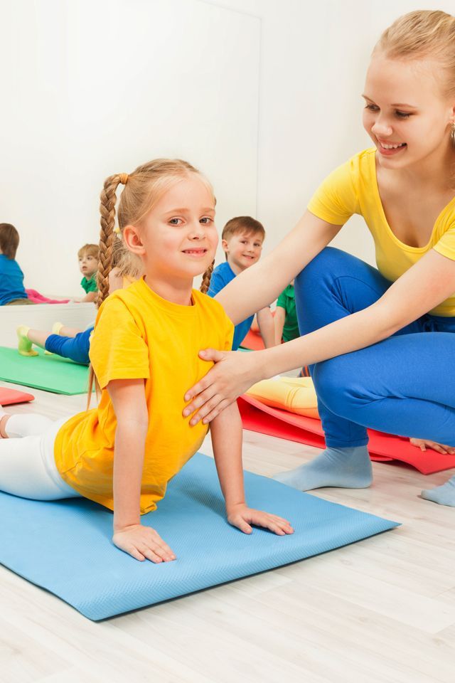 A girl doing a backbend on a blue mat, assisted by an instructor in a yellow shirt. Other children watch.