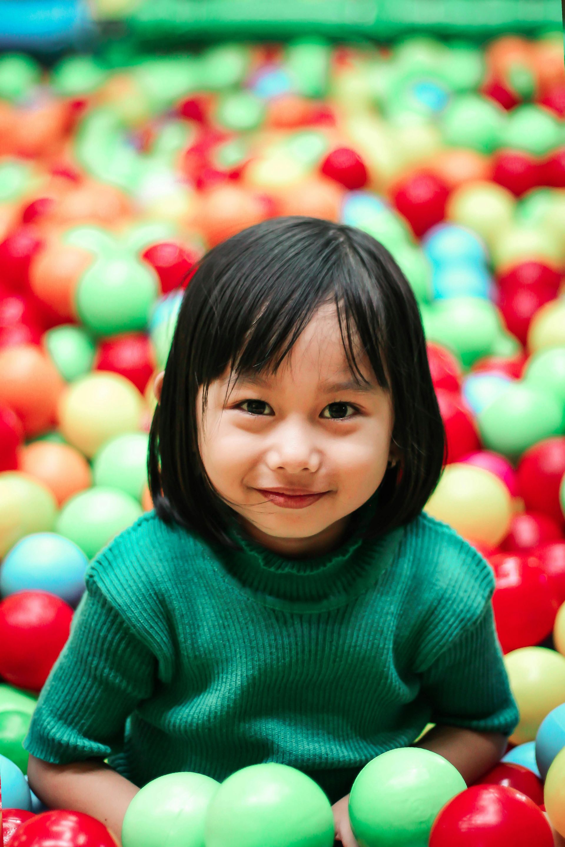 Child in a green sweater smiles, surrounded by a colorful ball pit.