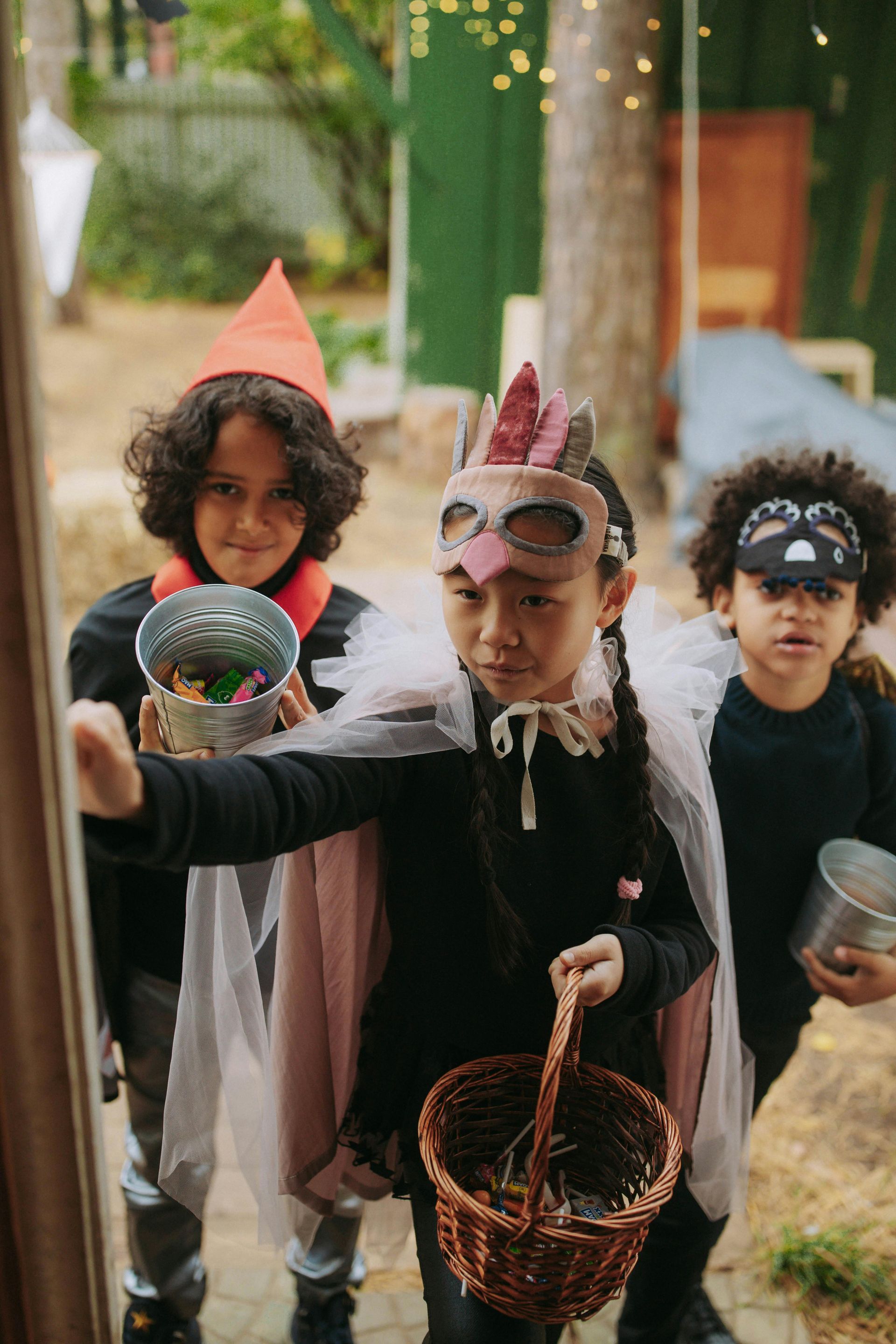 Three children in Halloween costumes trick-or-treating: a witch, a bird mask, and a cat.