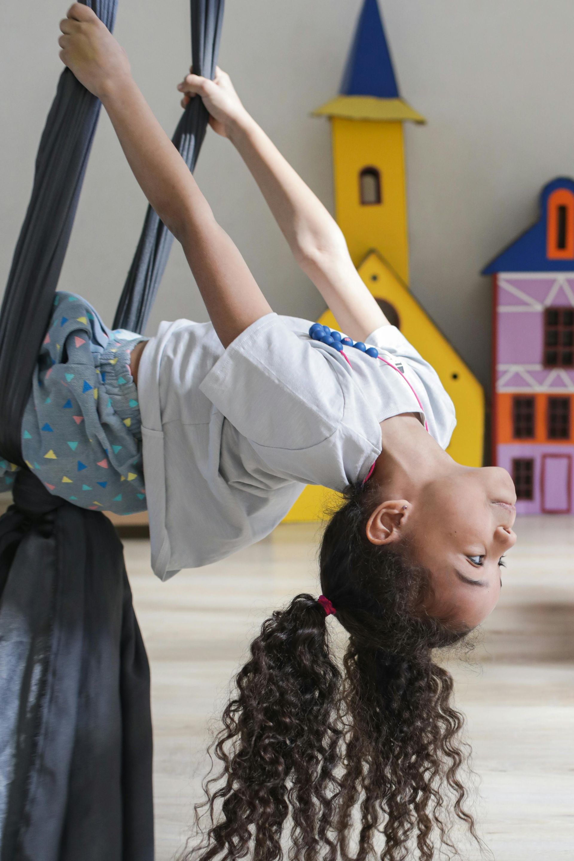 Girl upside down, holding aerial silks, smiling. Colorful toy buildings in background.