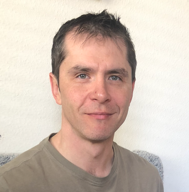 Man with short dark hair and light skin smiling slightly, wearing a tan shirt. White background.