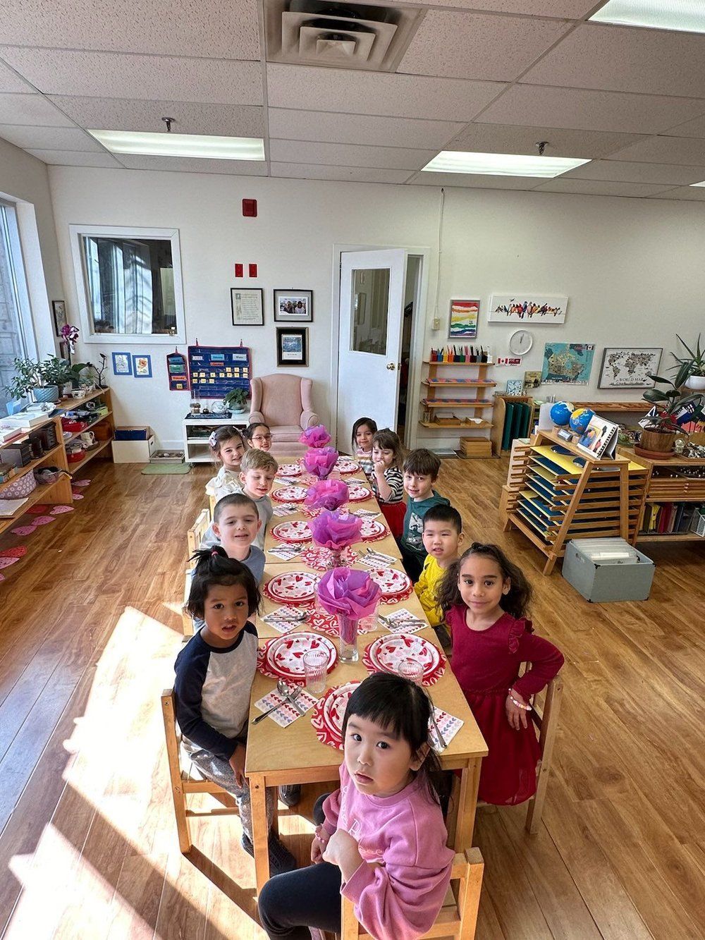A group of children are sitting at a long table in a classroom.