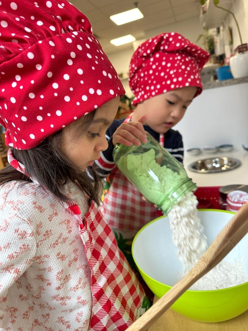Two children wearing chef hats are pouring ingredients into a bowl.