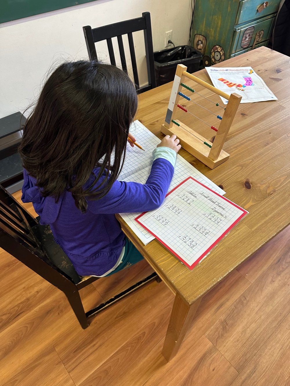 A little girl is sitting at a wooden table with a wooden abacus.