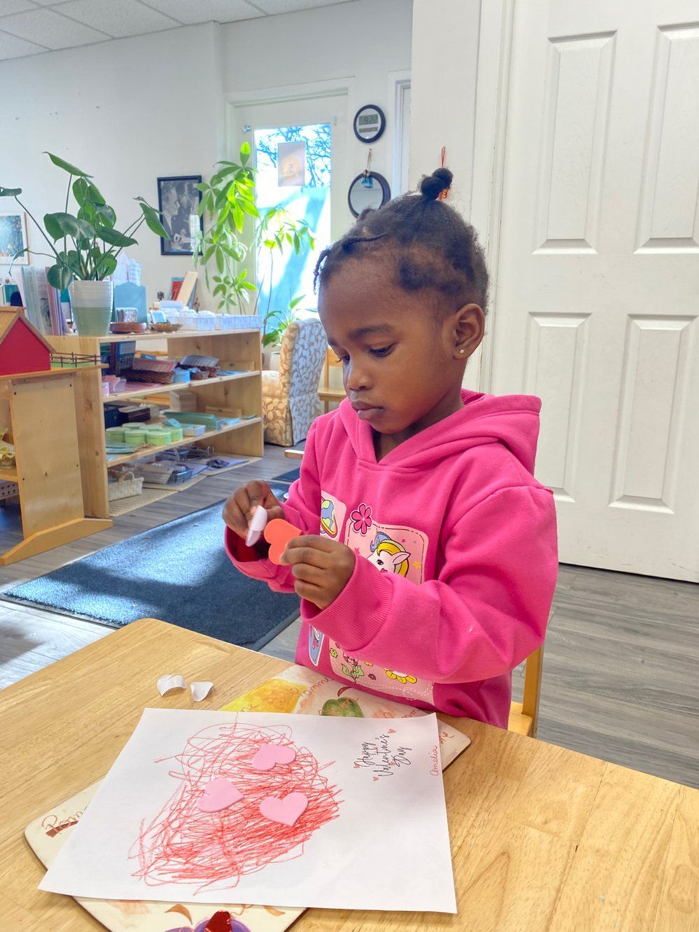 A little girl in a pink hoodie is sitting at a table playing with a red heart.