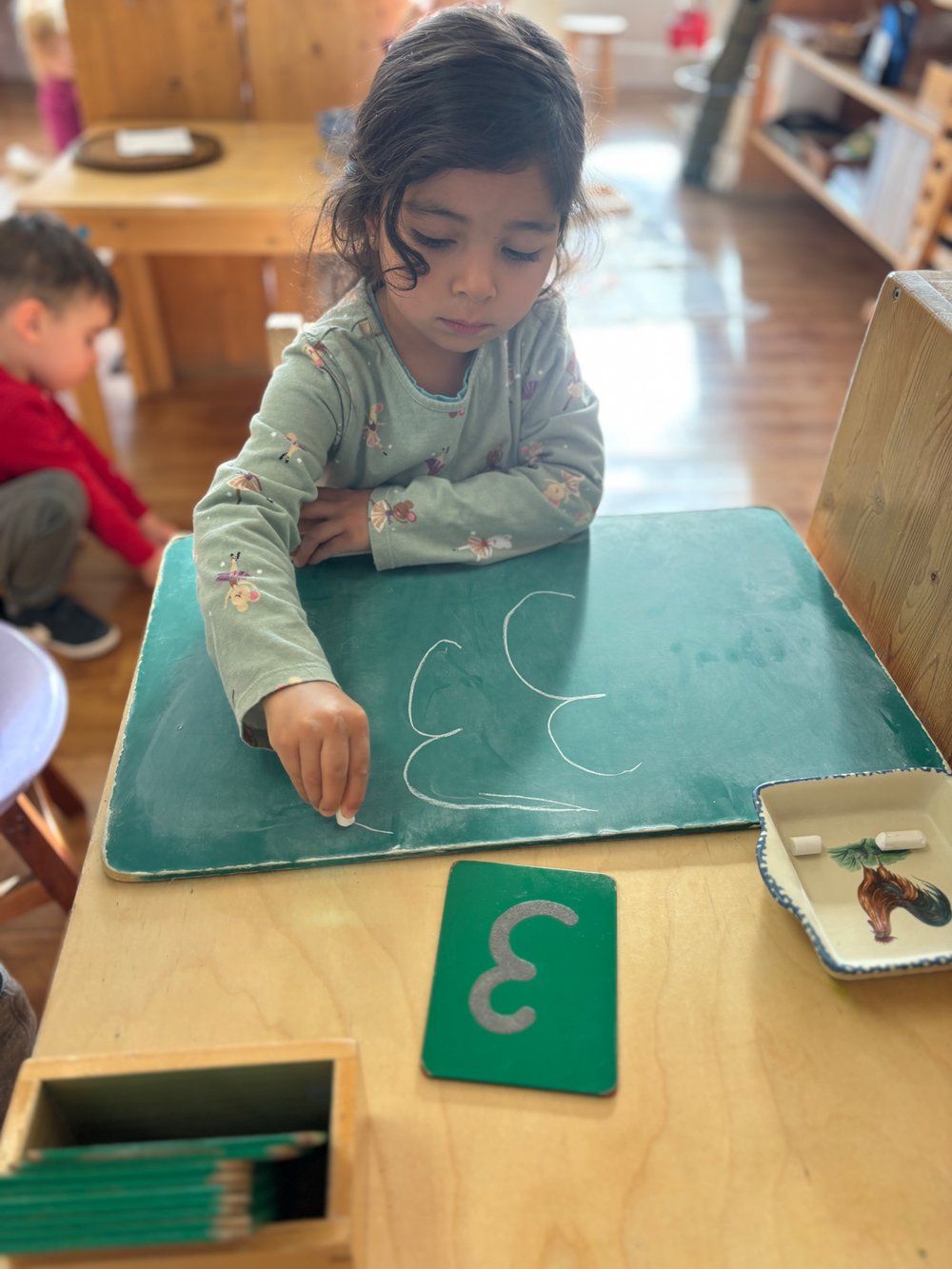 A little girl is sitting at a table writing on a chalkboard.