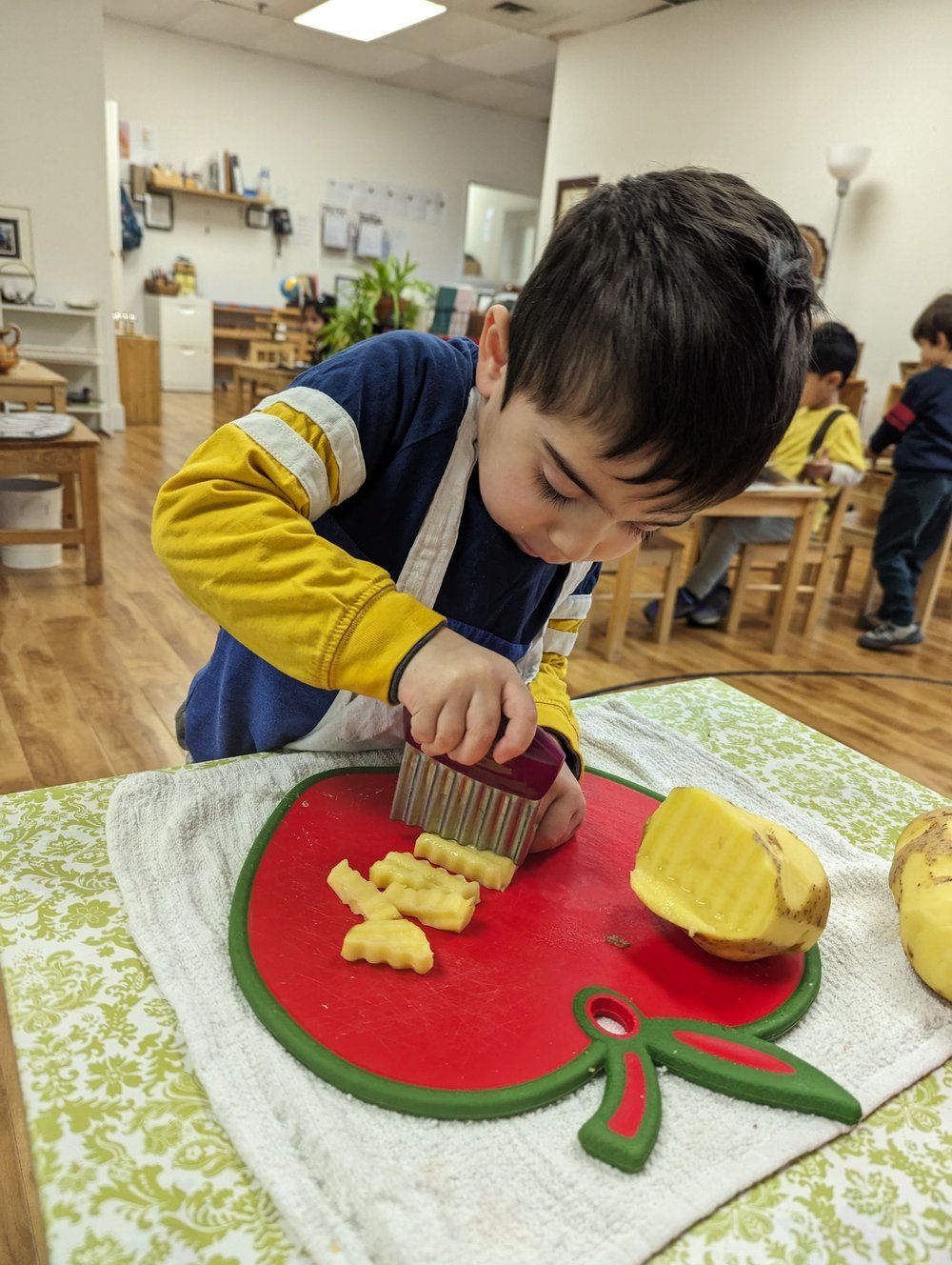 A young boy is cutting an apple on a cutting board in a classroom.