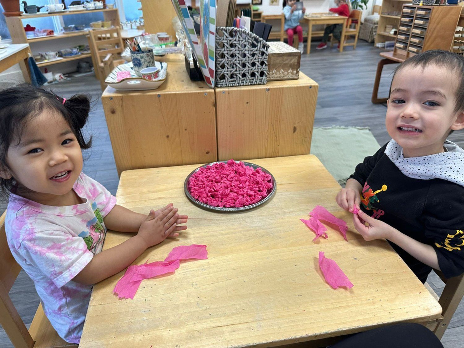 A boy and a girl are sitting at a table playing with pink tissue paper.