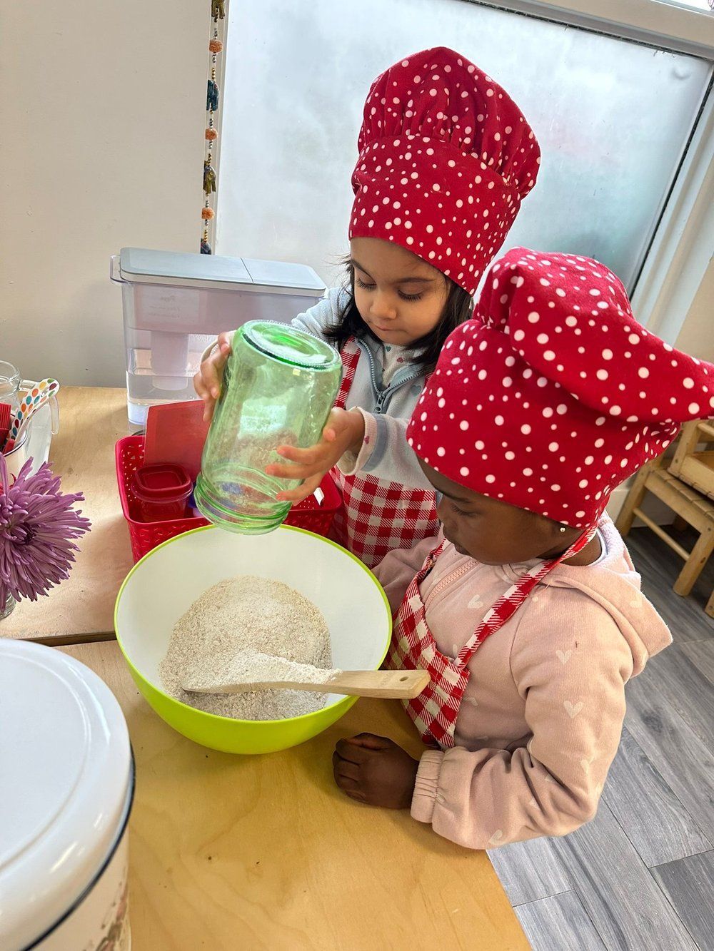 Two little girls wearing chef hats are pouring ingredients into a bowl.