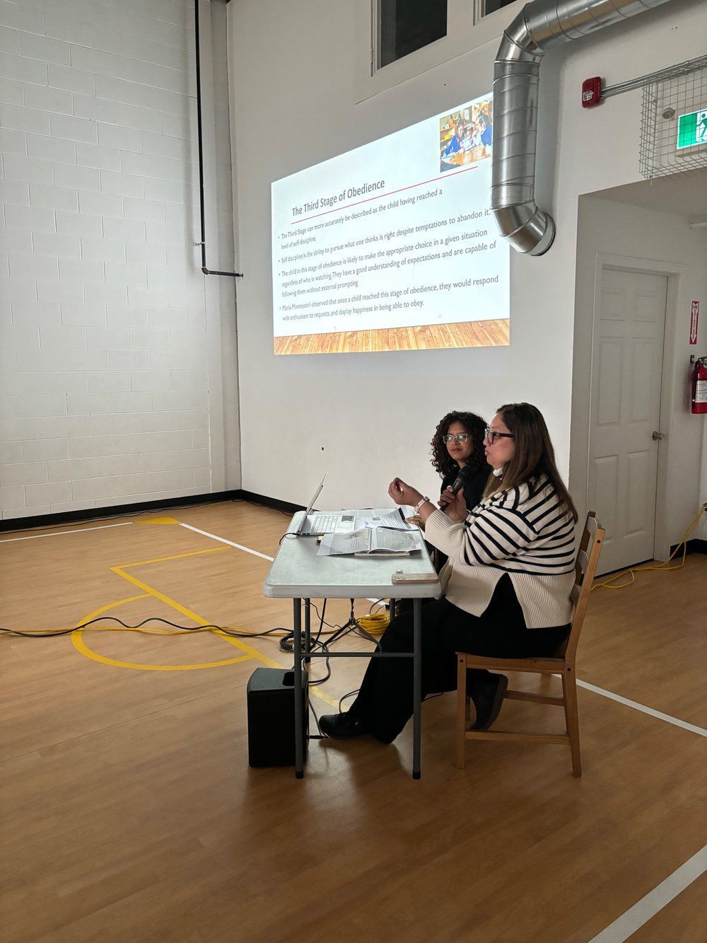 Two women are sitting at a table in front of a projector screen.