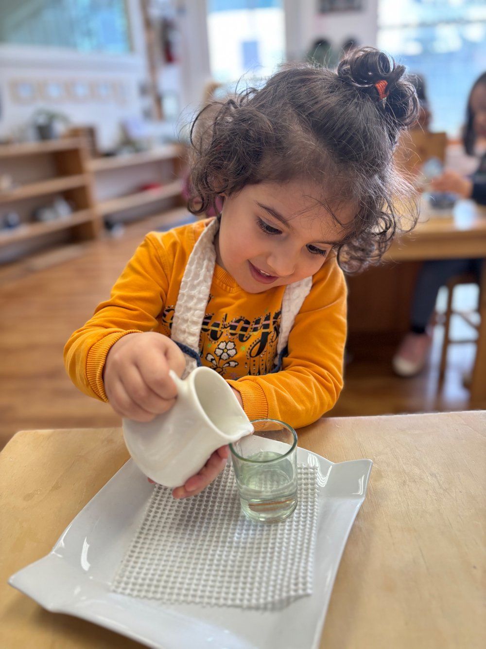 A little girl is pouring water into a glass on a plate.
