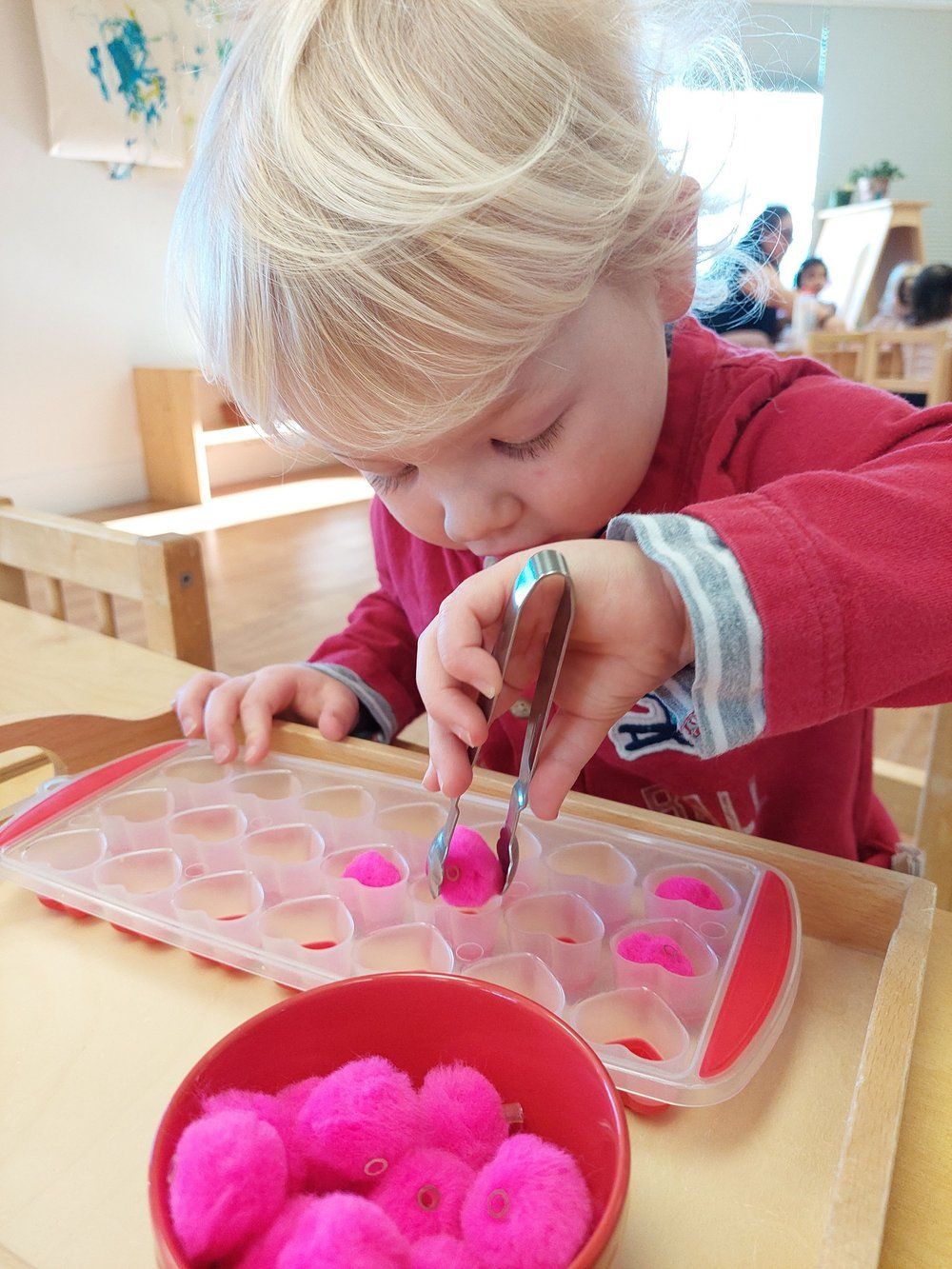 A little girl is playing with pink pom poms in an ice cube tray