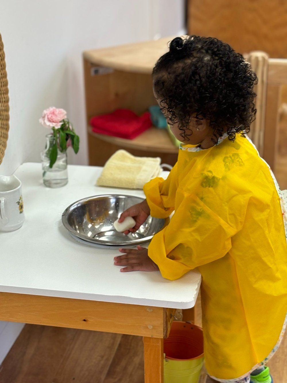 A little girl in a yellow apron is playing with a bowl on a table.