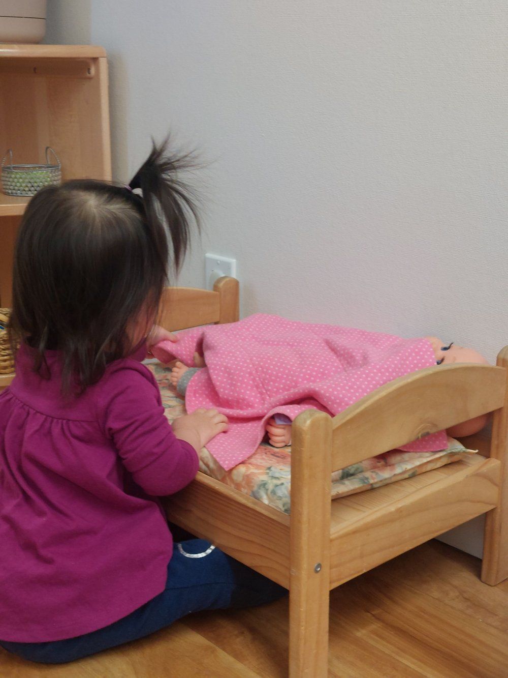 A little girl in a purple shirt sits on the floor next to a small wooden bed