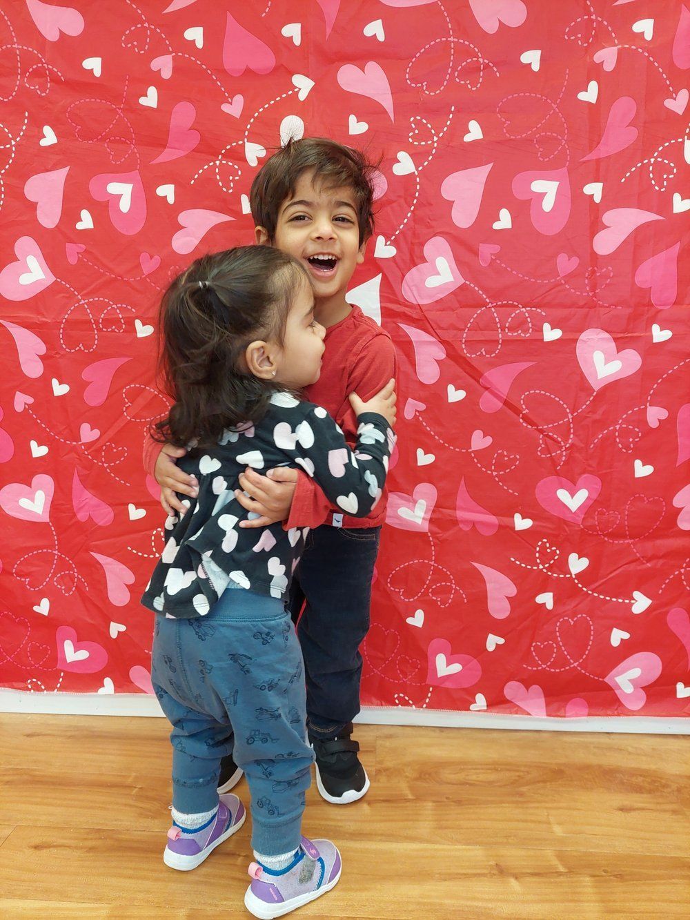A boy and a girl hugging in front of a wall with hearts on it