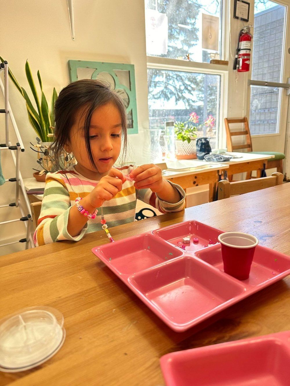 A little girl is sitting at a table with a pink tray and a red cup.