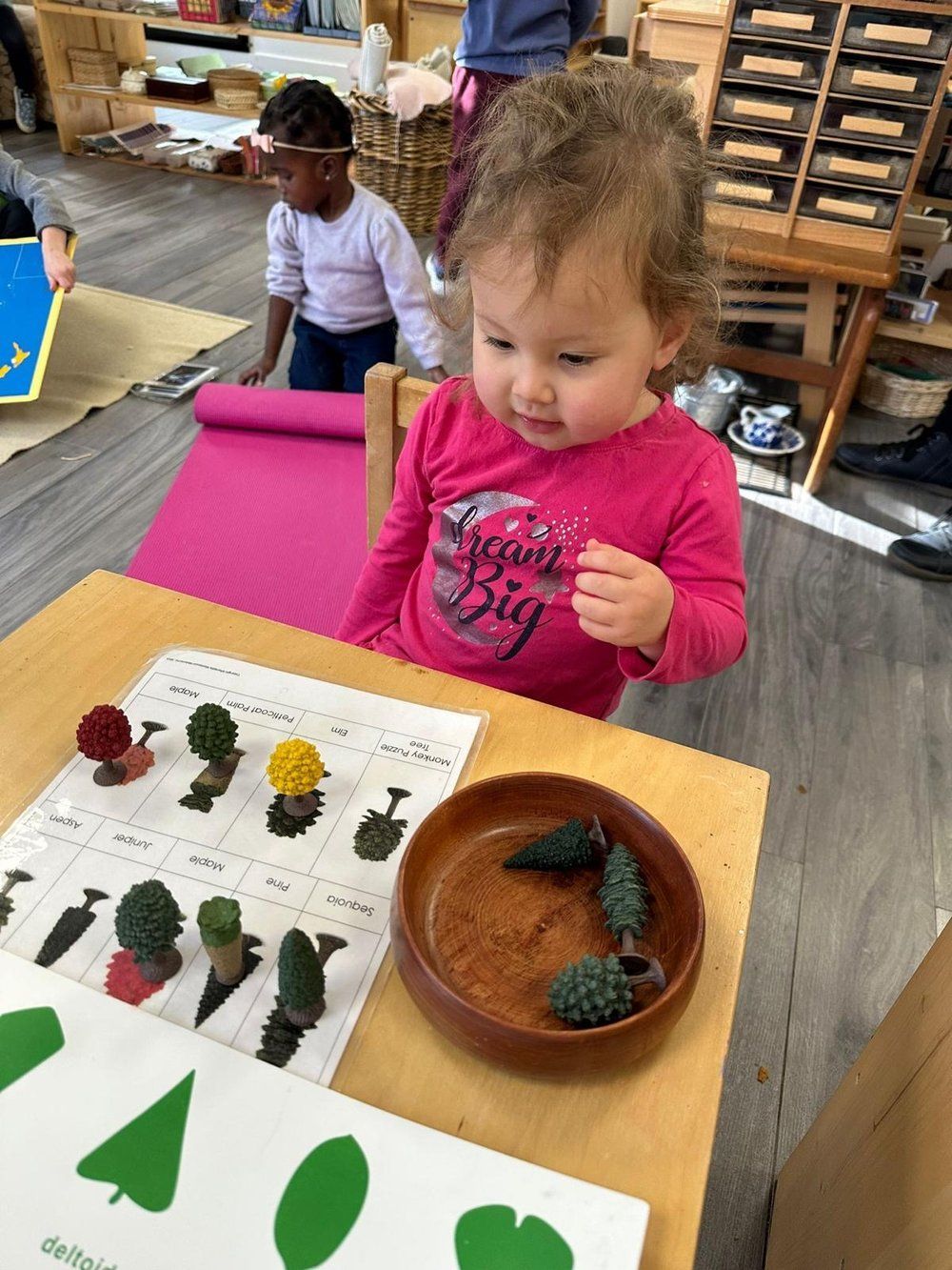 A little girl is sitting at a table playing with a bowl of plants.